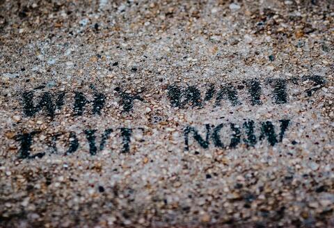 Image of stenciled text "WHY WAIT? LOVE NOW" in black paint on a gravelly surface