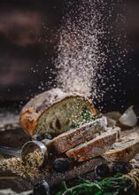 A photo of sliced bread with olives, rosemary, and cardamom pods, with breadcrumbs bursting into the air